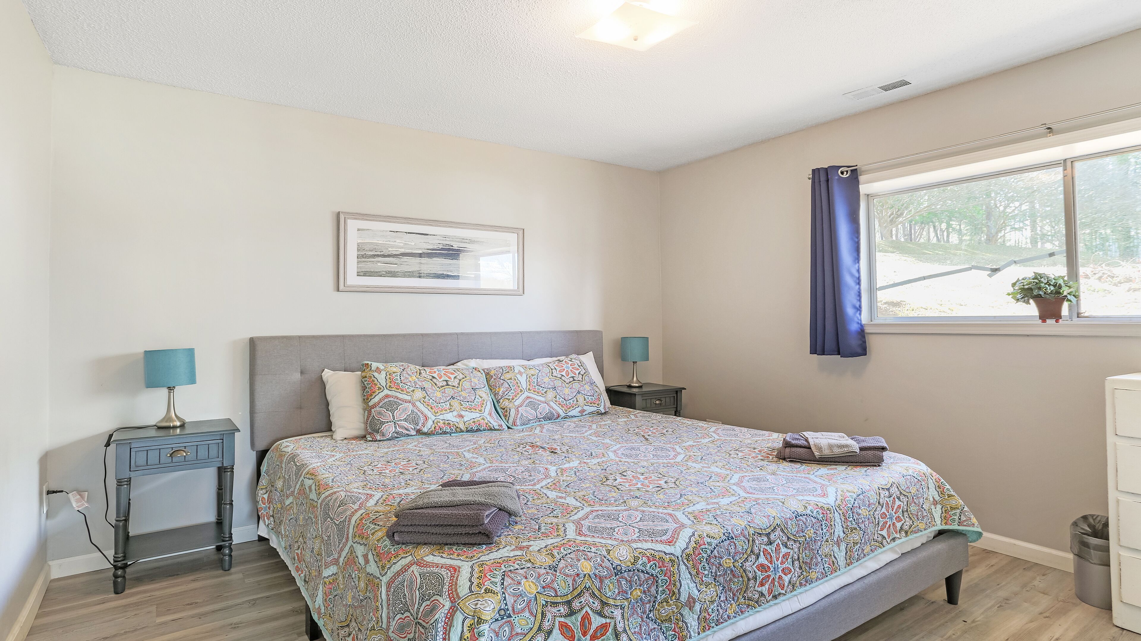 A bedroom with a large bed, two bedside tables with lamps, a window with a curtain, and a framed picture on the wall.