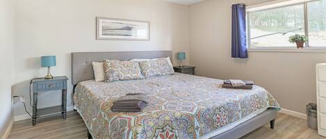 A bedroom with a large bed, two bedside tables with lamps, a window with a curtain, and a framed picture on the wall.