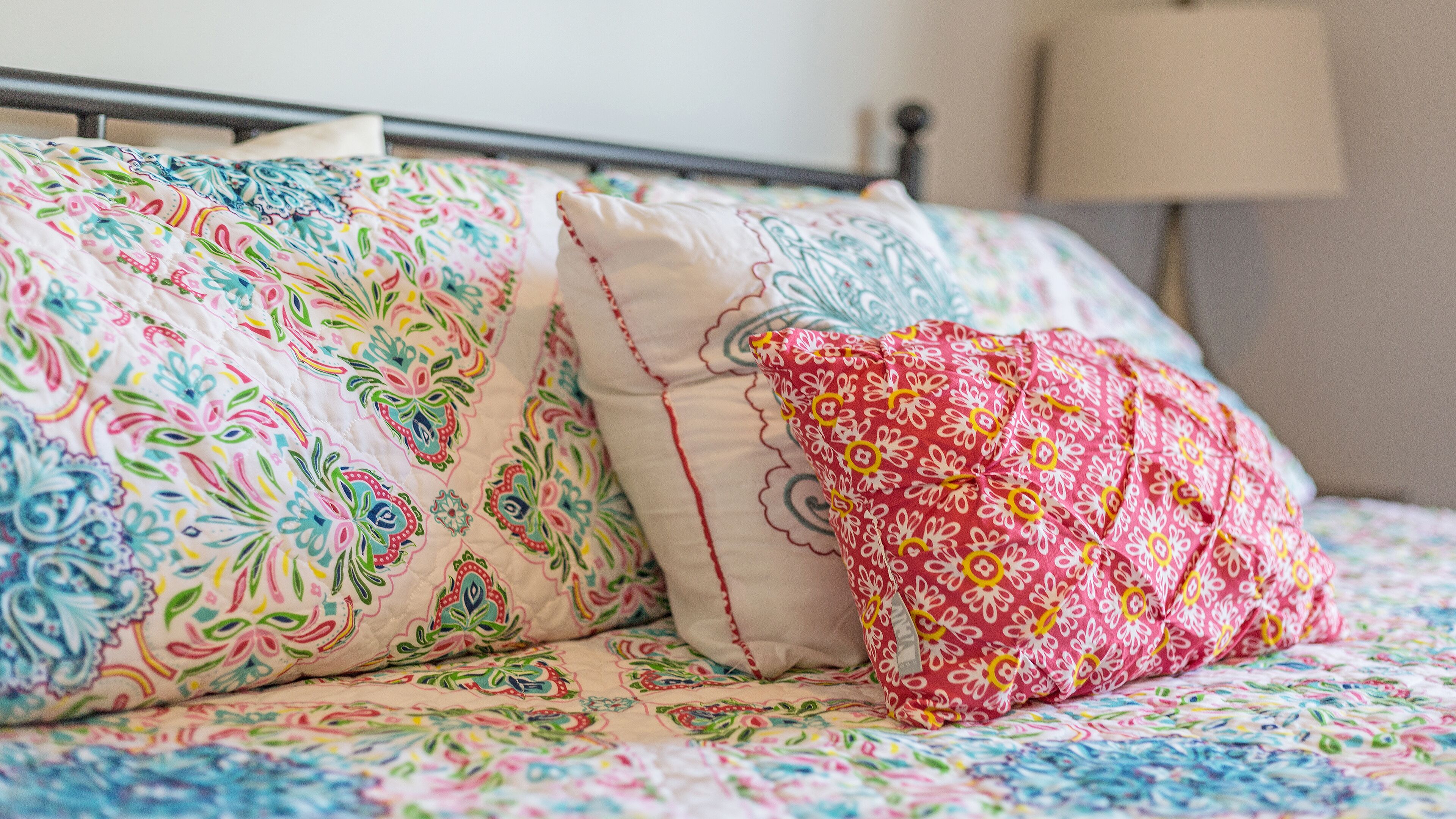 A bed with colorful floral bedding and two pillows, one with a paisley pattern and the other with a red and white floral design.