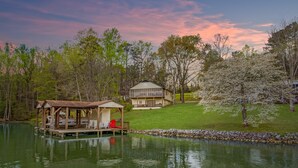 A lakeside cabin with a dock, a covered seating area, and a house in the background. - Lakefront 4bd-2ba; Deep water; Large Porch; Sleeps 14; Pet friendly (Macon)