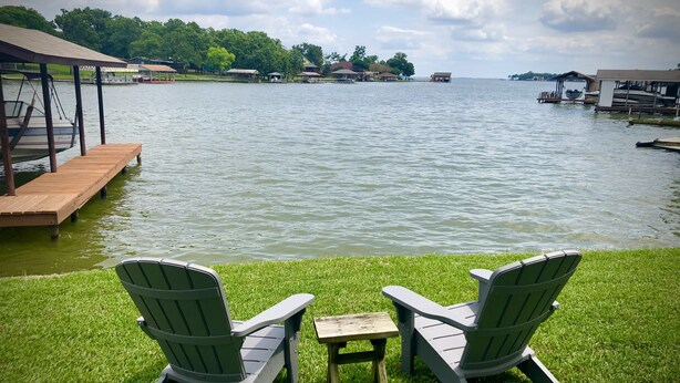 A lakeside view with two Adirondack chairs facing the water, a wooden dock, and a grassy area.