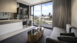 A modern hotel room with a kitchen area, a large window with a city view, and a seating area with a sofa and a coffee table. - Majestic M Suites (North Adelaide)