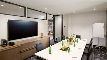 A conference room with a long table, chairs, a large flat-screen TV, a whiteboard, and a wooden cabinet with a coffee machine.