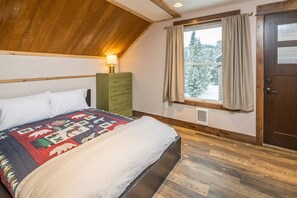 Ein Schlafzimmer mit Holzboden, einem Bett mit Patchworkdecke, einer grünen Kommode, einem Fenster mit Vorhängen und einer Tür.