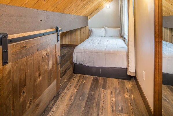 A bedroom with a wooden slat ceiling, a bed with white bedding, and a wooden floor.