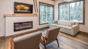 Ein gemütliches Wohnzimmer mit Kamin, großen Fenstern mit Blick auf einen verschneiten Wald, einer Couch und einem Fernseher, der über dem Kamin angebracht ist.