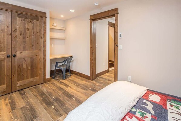 A bedroom with a wooden wardrobe, a desk with a chair, and a bed with a patterned blanket.