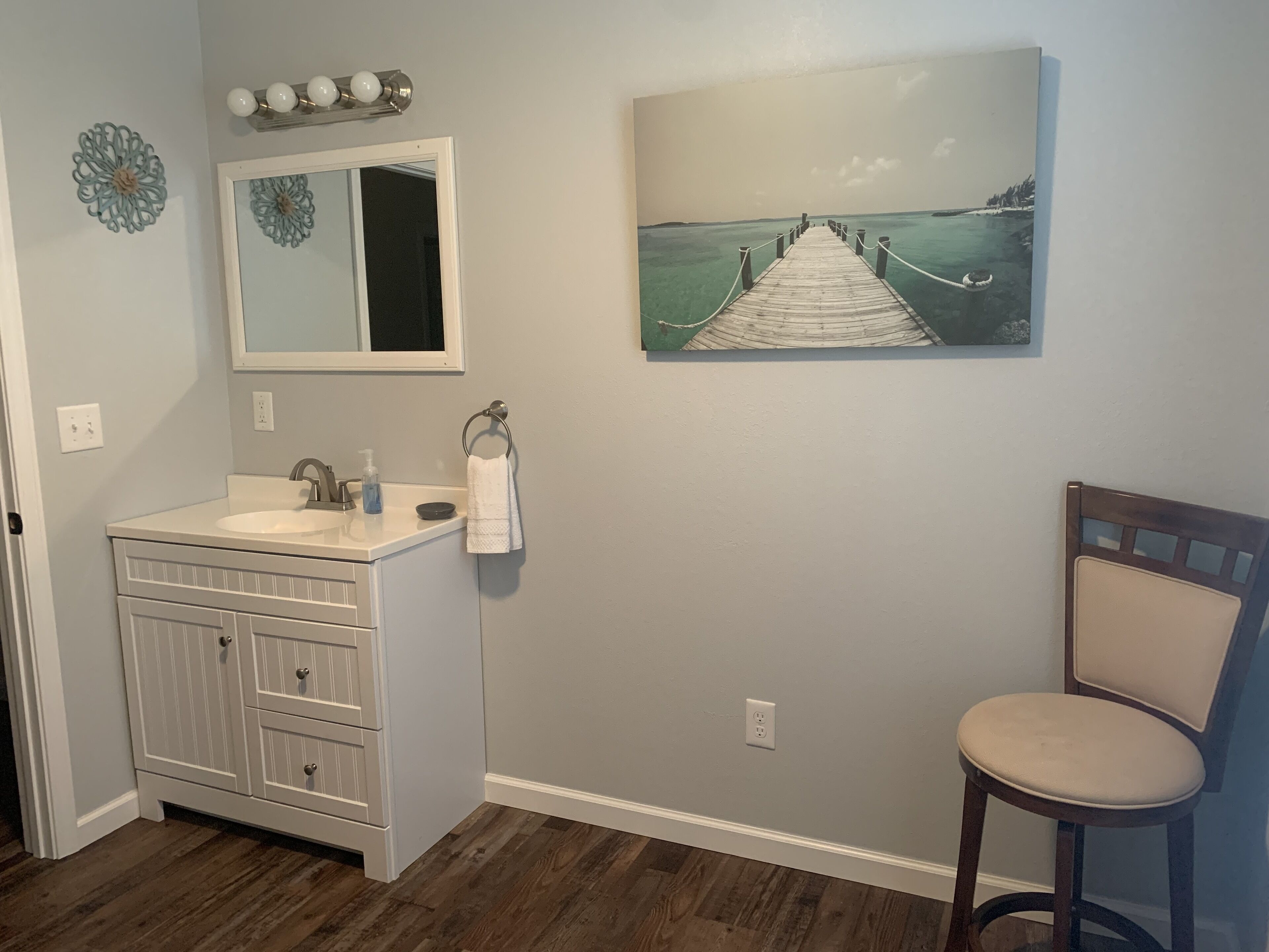 Un baño con tocador blanco, espejo, pintura de un muelle y una silla de madera.