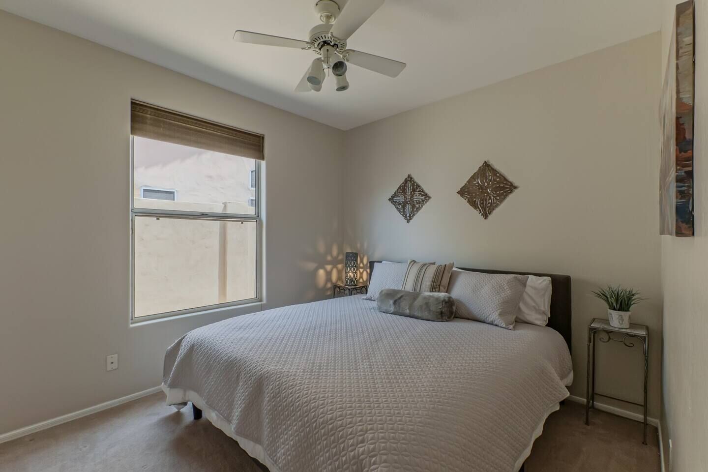 A bedroom with a large bed, a ceiling fan, a window with blinds, decorative wall art, and a small side table with a plant.