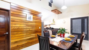 A dining area with a wooden wall, a table set for four, and a kitchen in the background. - In the middle of the best beaches, private house with pool. (Tamarindo)