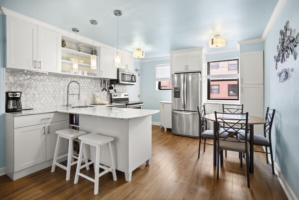 Cocina moderna con armarios blancos, una isla central con taburetes, electrodomésticos de acero inoxidable y una zona de comedor con una mesa redonda y sillas.