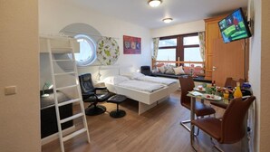 Habitación pequeña con literas, un escritorio y silla, una mesita con otra silla, televisor montado en la pared y ventana con cortinas.