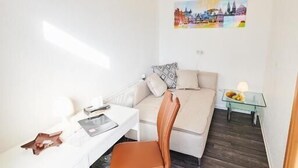 Una habitación compacta con un escritorio blanco, una silla marrón, un sofá y una mesita de noche con lámpara.