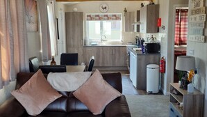 A living room with a brown leather sofa, a dining area with a table and chairs, and a kitchen with wooden cabinets and a window. - Captivatingly Stunning 2-bed Chalet in Bridlington (Bridlington)
