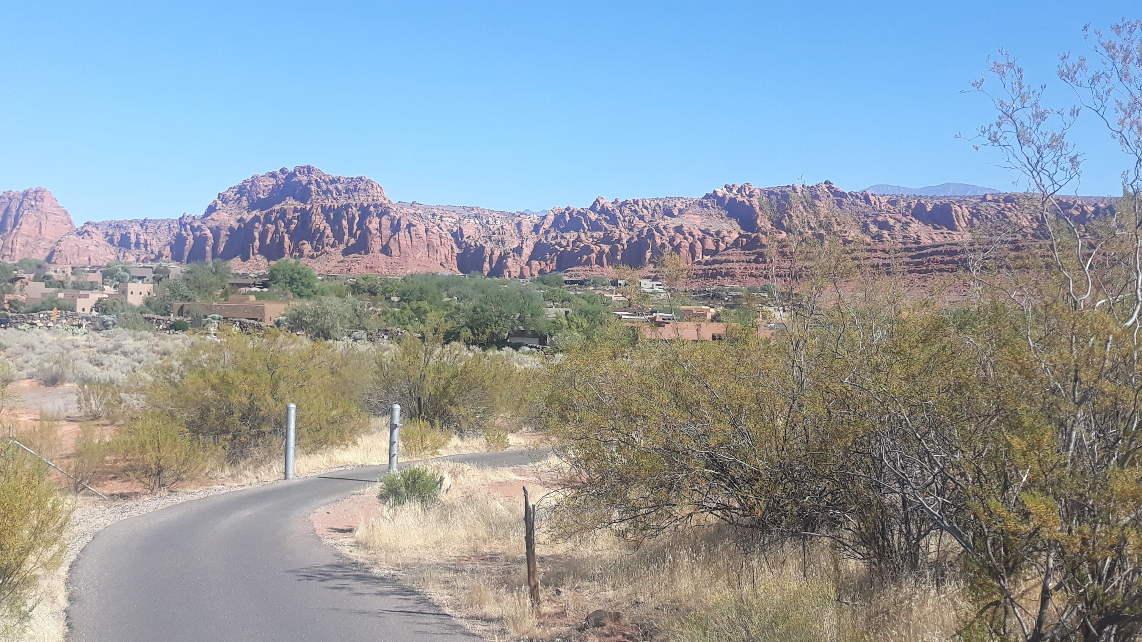 A road curving through a semi-arid landscape with sparse vegetation and distant red rock formations.