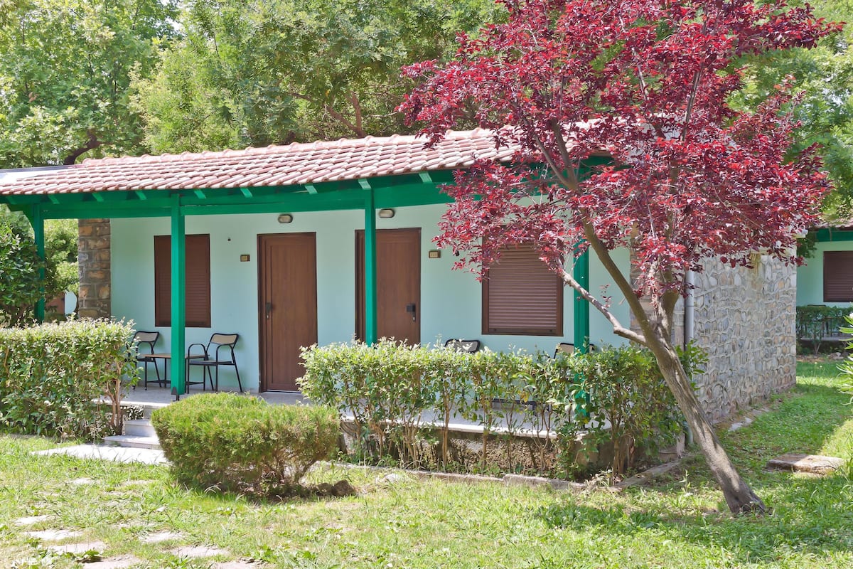 Un petit bâtiment doté d’un toit en tuiles, de piliers verts et d’un petit jardin agrémenté d’un arbre et d’arbustes.