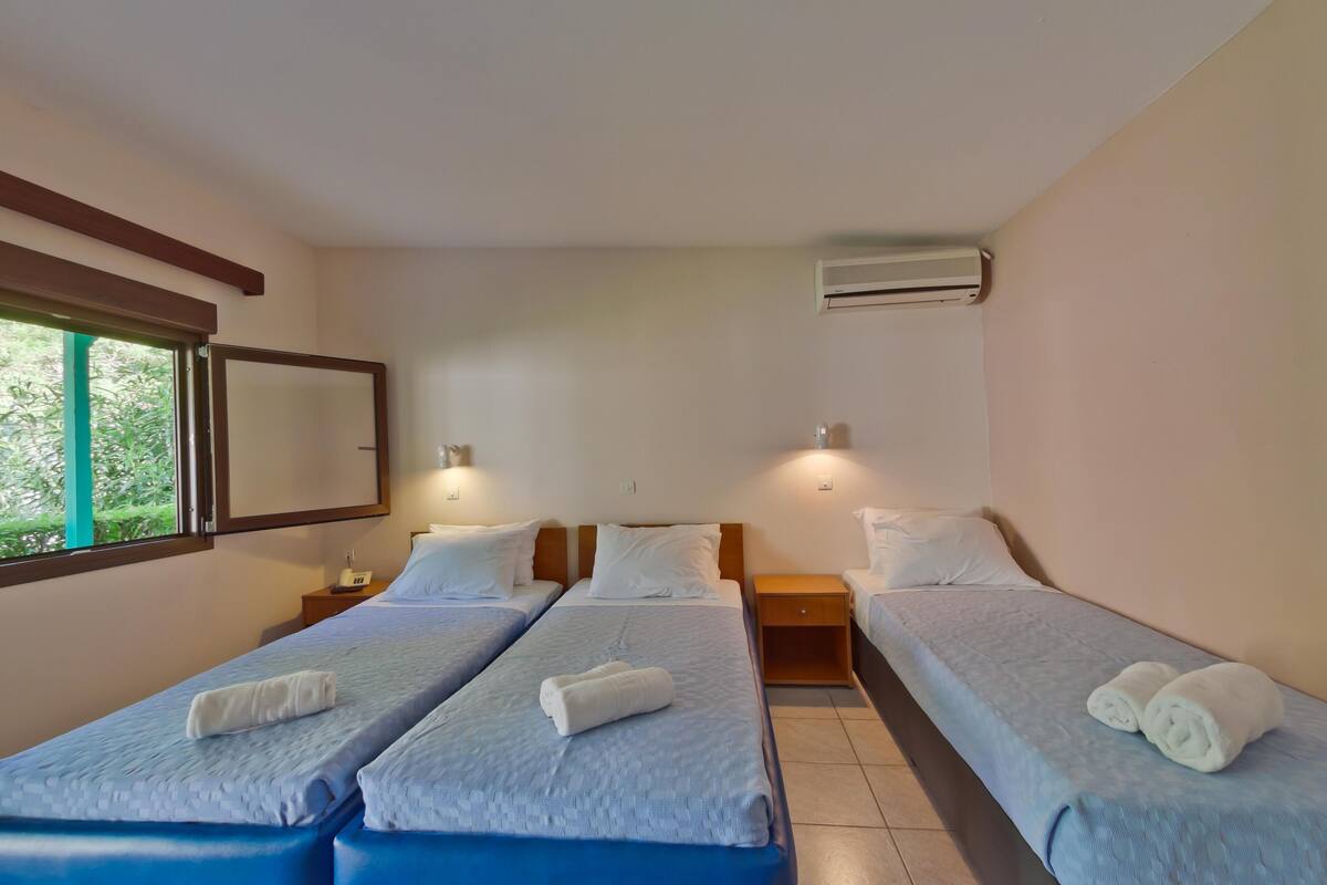 Une chambre d’hôtel avec trois lits simples, chacun recouvert d’une couverture bleue et agrémenté d’oreillers blancs. On y trouve une table de chevet en bois avec une lampe, placée entre les lits, ainsi qu’un appareil de climatisation fixé au mur.