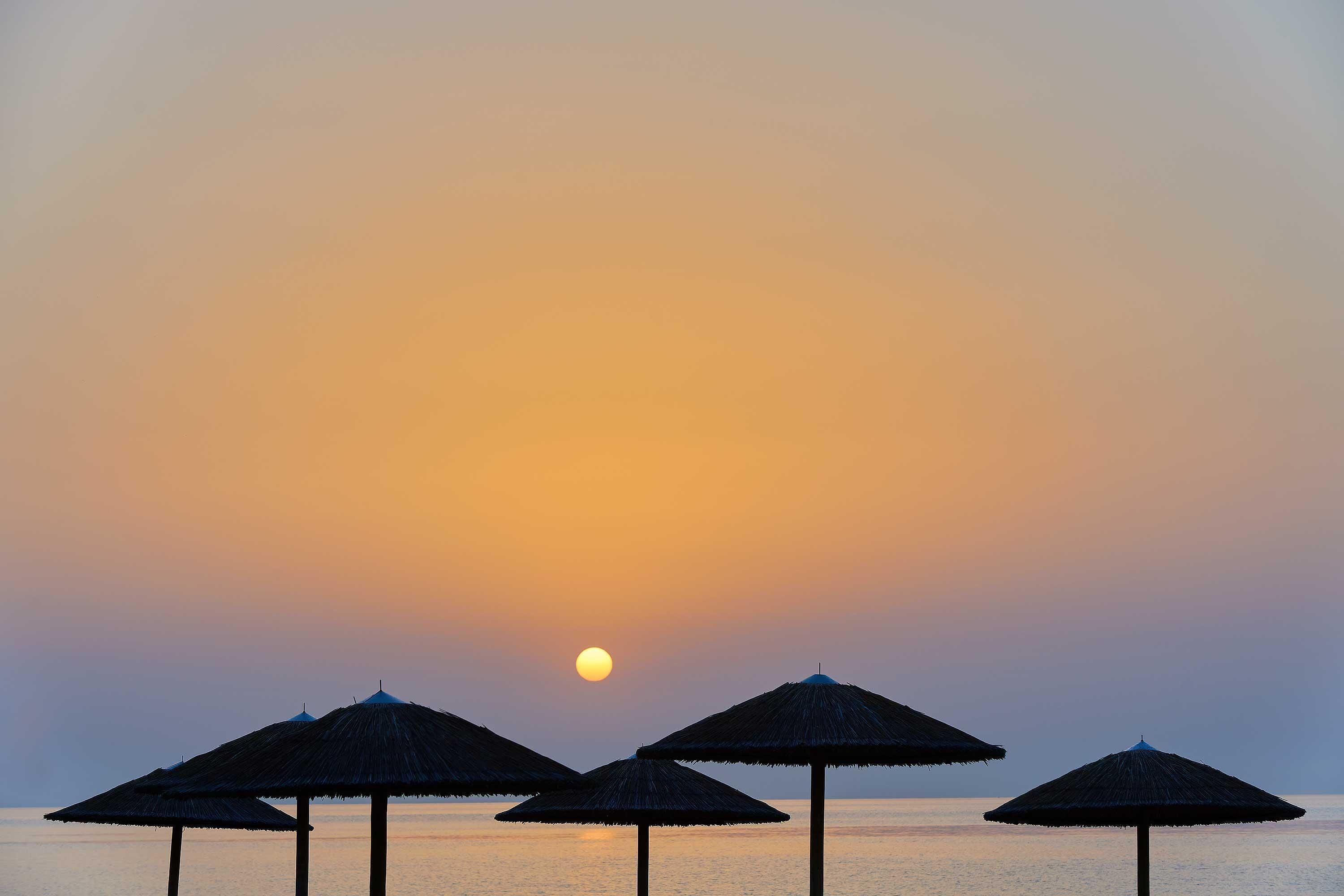 A sunset over a calm sea with three thatched umbrellas.