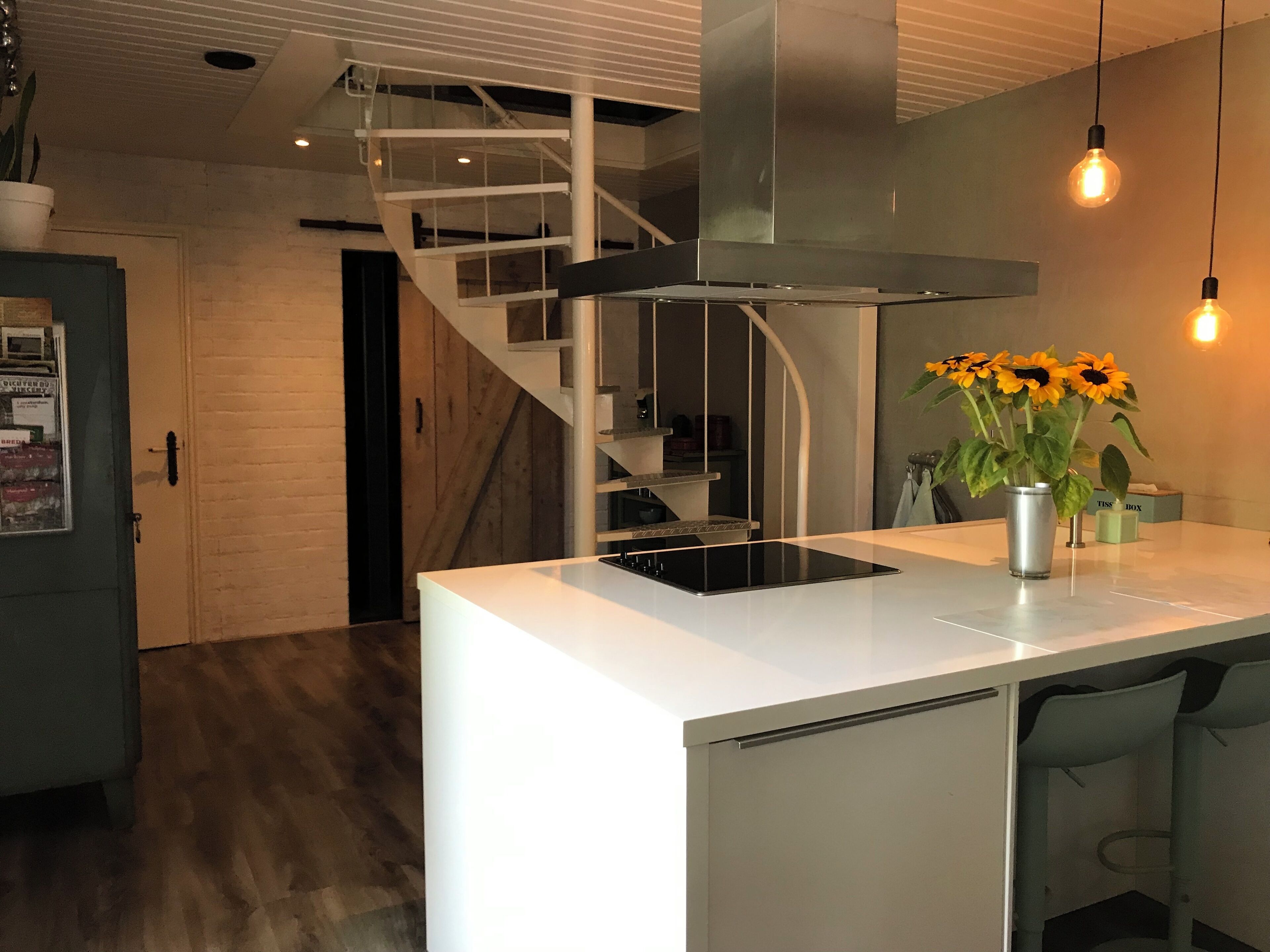 A modern kitchen with a central island, a stainless steel range hood, a staircase, and hanging pendant lights.