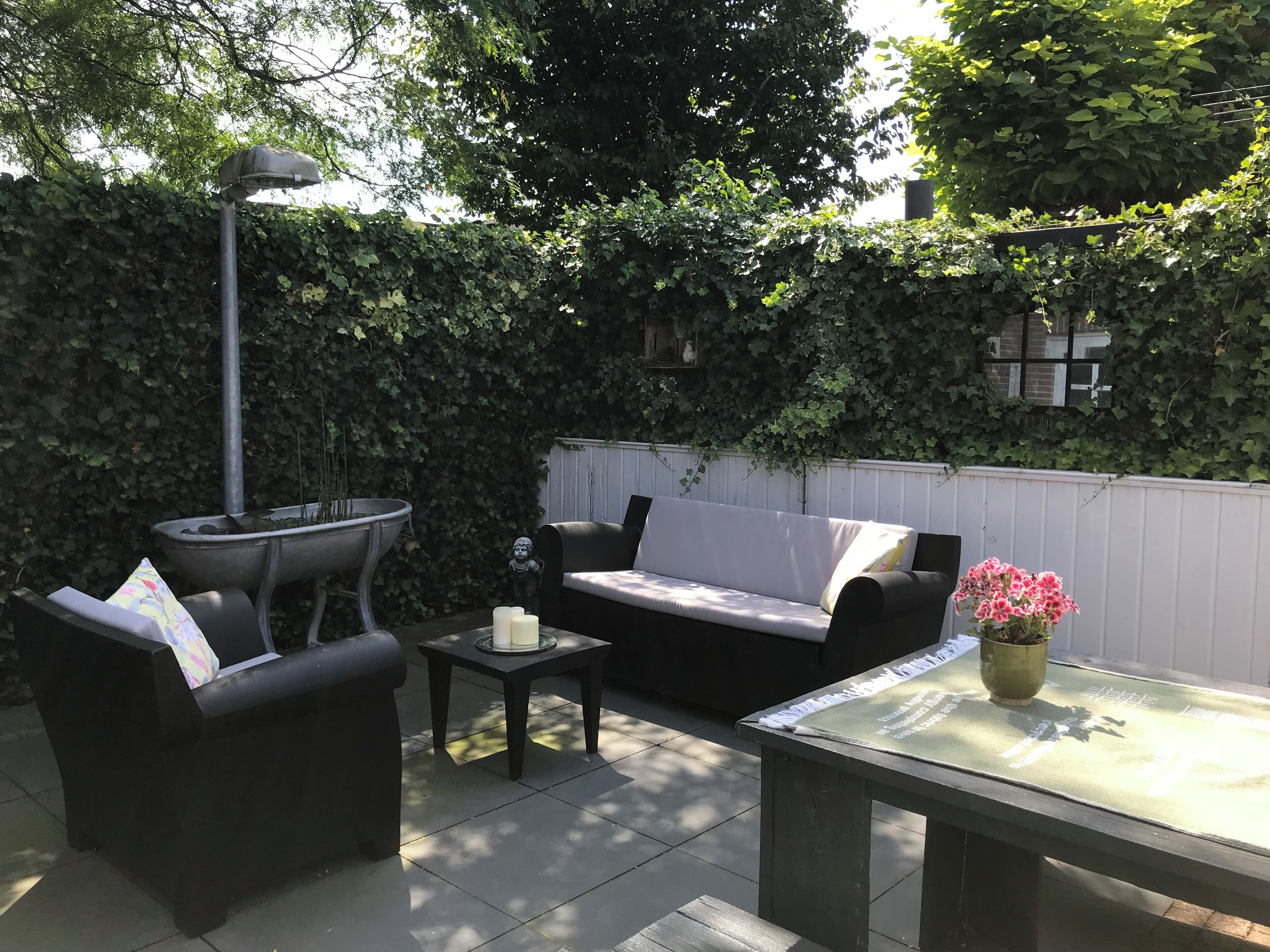 A patio area with a black sofa, a small table, and a chair. There is a decorative lamp and a potted plant with pink flowers.