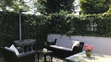 A patio area with a black sofa, a small table, and a chair. There is a decorative lamp and a potted plant with pink flowers.