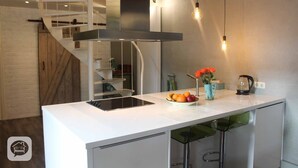 Una cocina moderna con encimera blanca, horno empotrado, campana extractora de acero inoxidable y un frutero con naranjas y manzanas.