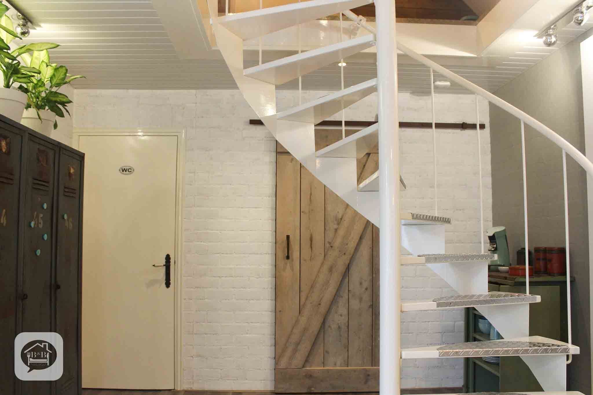 A spiral staircase with a white railing, a wooden door with a 'WC' sign, and a plant in the corner.
