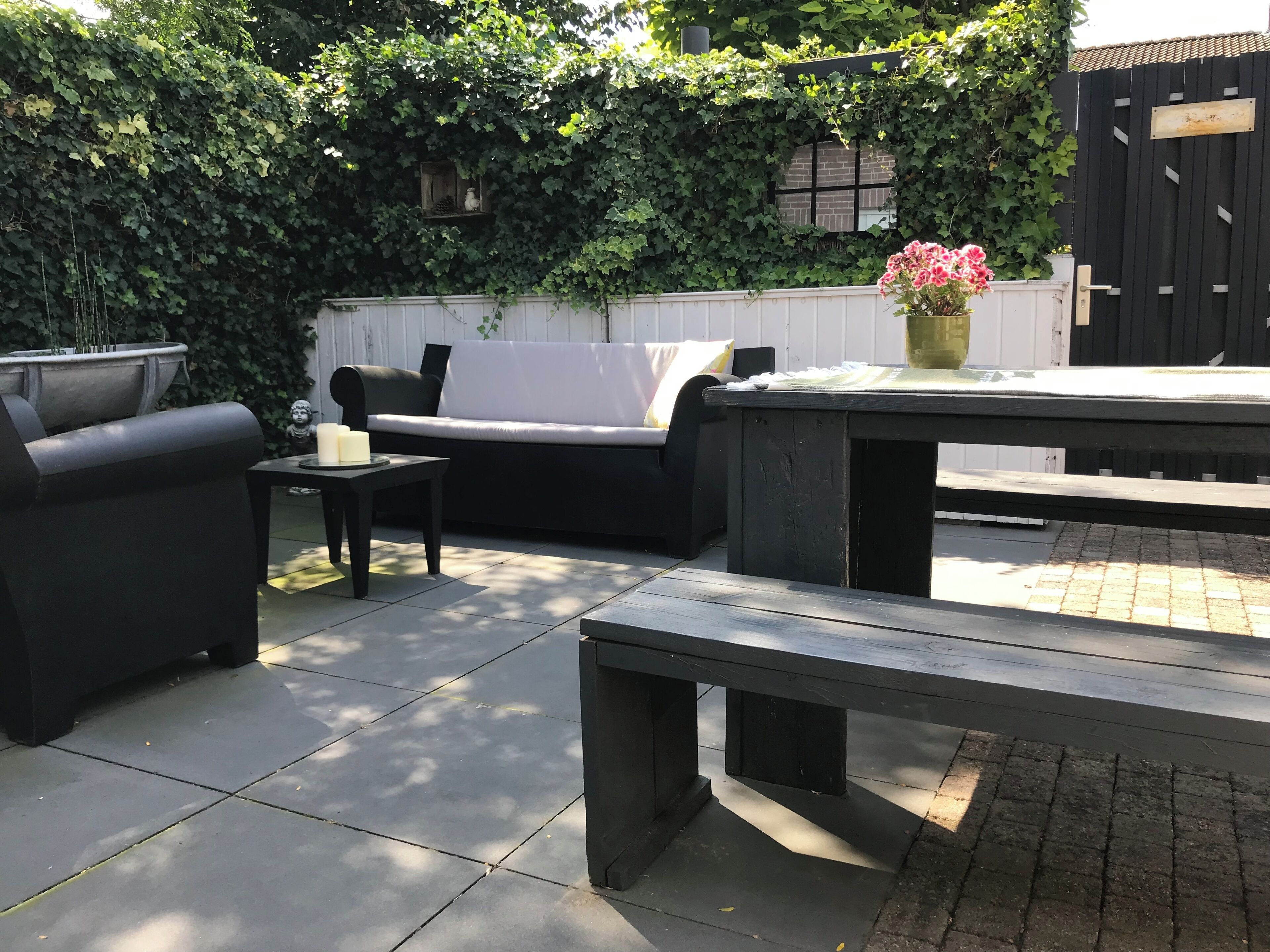 Un espace extérieur aménagé en terrasse, avec une table noire, un canapé noir et un fauteuil noir. On y trouve une petite table avec des bougies et un vase rempli de fleurs. La terrasse est entourée d’une clôture blanche et d’un portail noir.