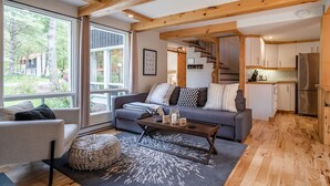 A modern living room with a sofa, coffee table, and ottoman, wooden beams, large windows, and a kitchen area with cabinets and a refrigerator. - Chalet Scandinavian,  Turn Key Amenities + Pet Friendly + Dedicated Workspace with Hi Speed WiF (Orford)