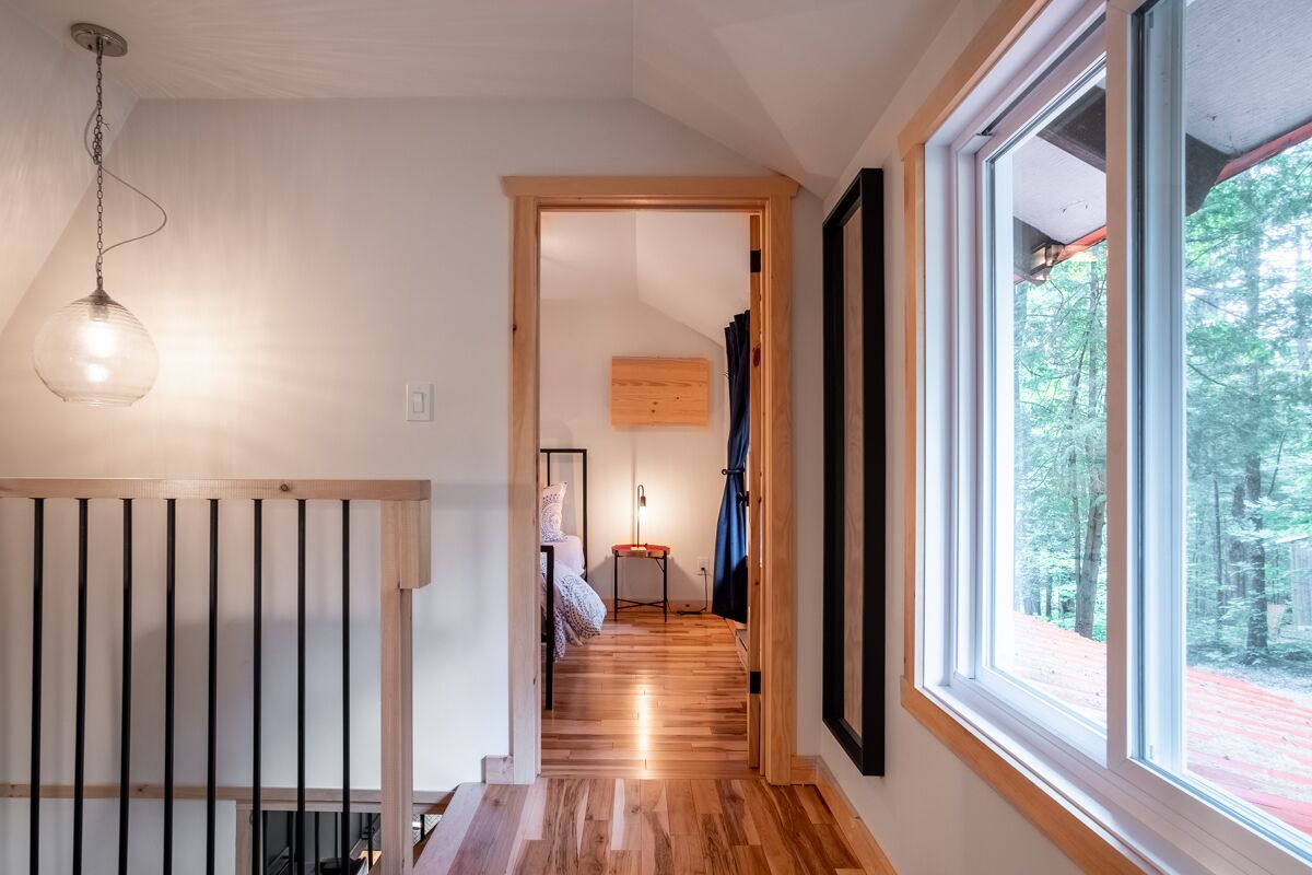A hallway with wooden flooring, a hanging light fixture, and a large window with a view of trees.