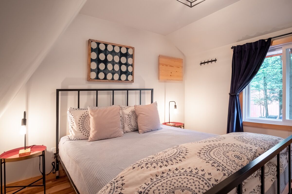 A bedroom with a sloped ceiling, a bed with patterned bedding, a bedside table with a lamp, and a window with curtains.