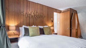 Habitació d'hotel amb paret de panells de fusta, un llit amb roba de llit blanca i vàries almoadines, una meseta de nit amb una làmpada i un armari.