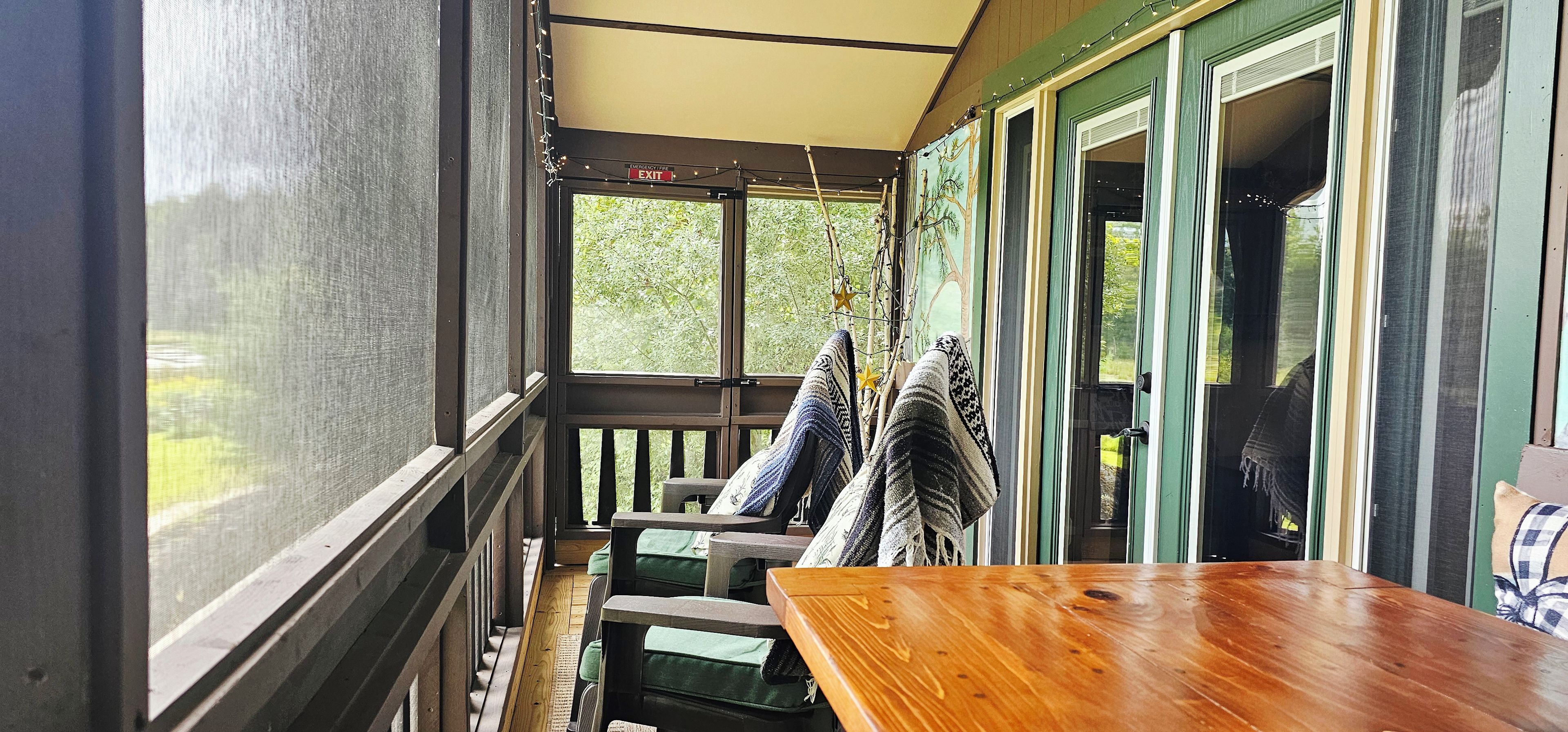 Een overdekt terras met een houten tafel en stoelen, uitzicht op groen en een deur met een nooduitgangsbord.