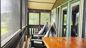 Een overdekt terras met een houten tafel en stoelen, uitzicht op groen en een deur met een nooduitgangsbord.