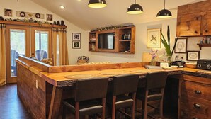 A wooden bar area with a counter, bar stools, a television, and a kitchen area with wooden cabinets and a shelf. - ADK Rustic Private Chalet Viewing Two Ponds / Forest 5.3 miles To Gore Mountain (North Creek)