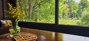 A window with a view of a forest and a wooden table with a vase of flowers and a tray of food. - ADK Rustic Private Chalet Viewing Two Ponds / Forest 5.3 miles To Gore Mountain (North Creek)
