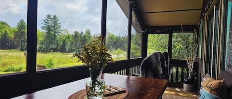 A room with a large window overlooking a forested area, a wooden table with a vase of flowers, and a chair.