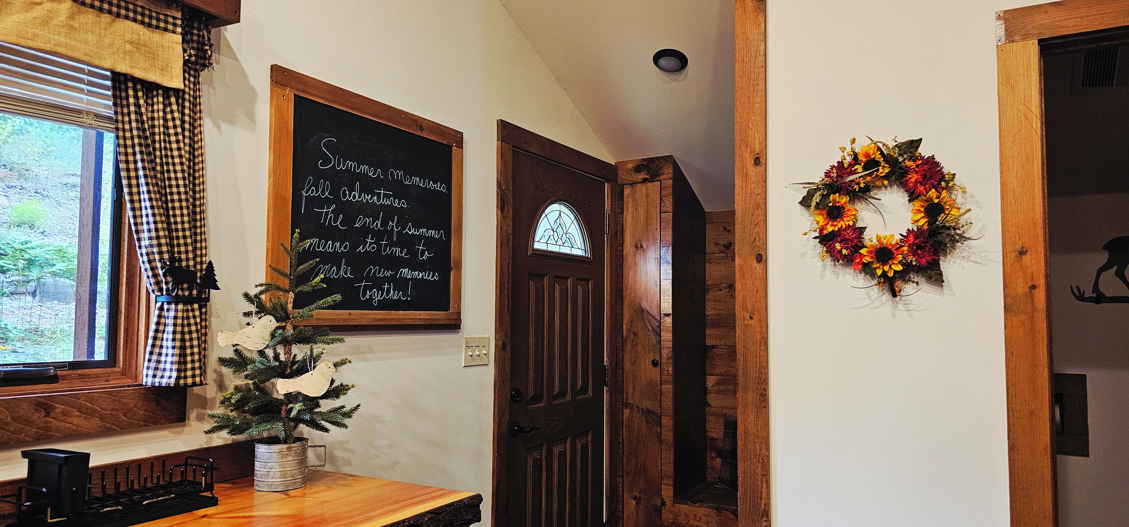 A room with a wooden door, a small Christmas tree, a chalkboard with a message, and a wreath on the wall.