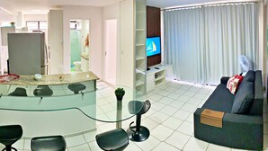 A modern living room with a sofa, a TV, a dining area with a glass table and chairs, and a kitchen in the background. - Vacanze - Monteverdi (Maceió)
