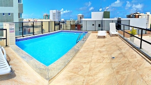 En pool på taget med klart blåt vand, omgivet af en flisbelagt terrasse og hvide liggestole.
