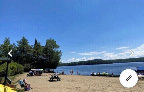 A lakeside beach with picnic tables, umbrellas, and boats.