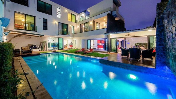 Una casa moderna de dos pisos con piscina, mobiliario de exterior e interior bien iluminado.