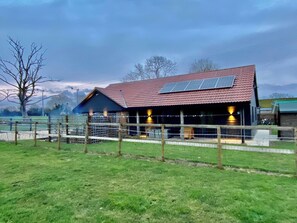 Een schuur met een rood dak en zonnepanelen, omgeven door een omheind gebied met een grasveld.