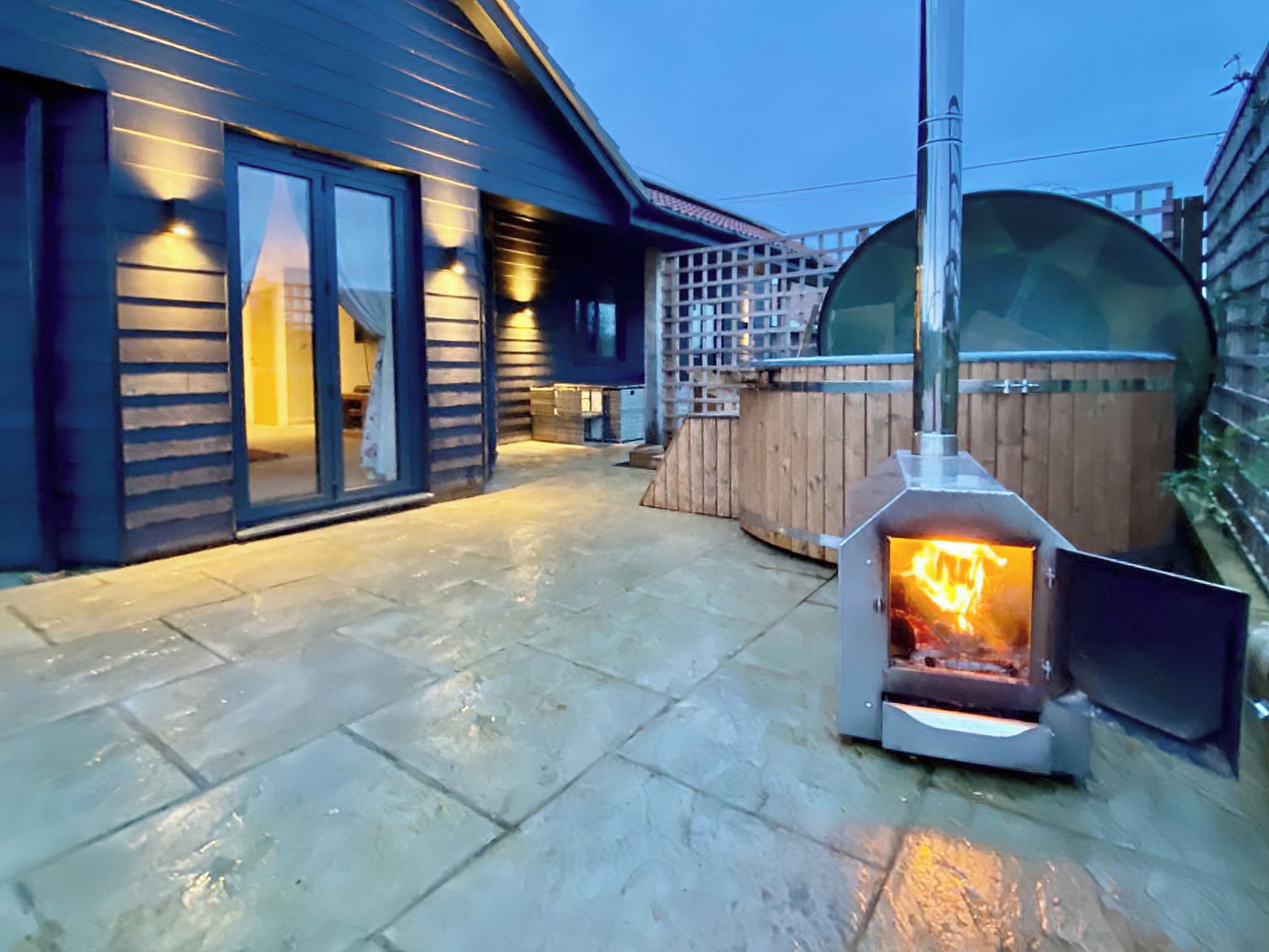 Une terrasse extérieure avec un poêle à bois, une clôture en bois et un bâtiment doté de grandes fenêtres.
