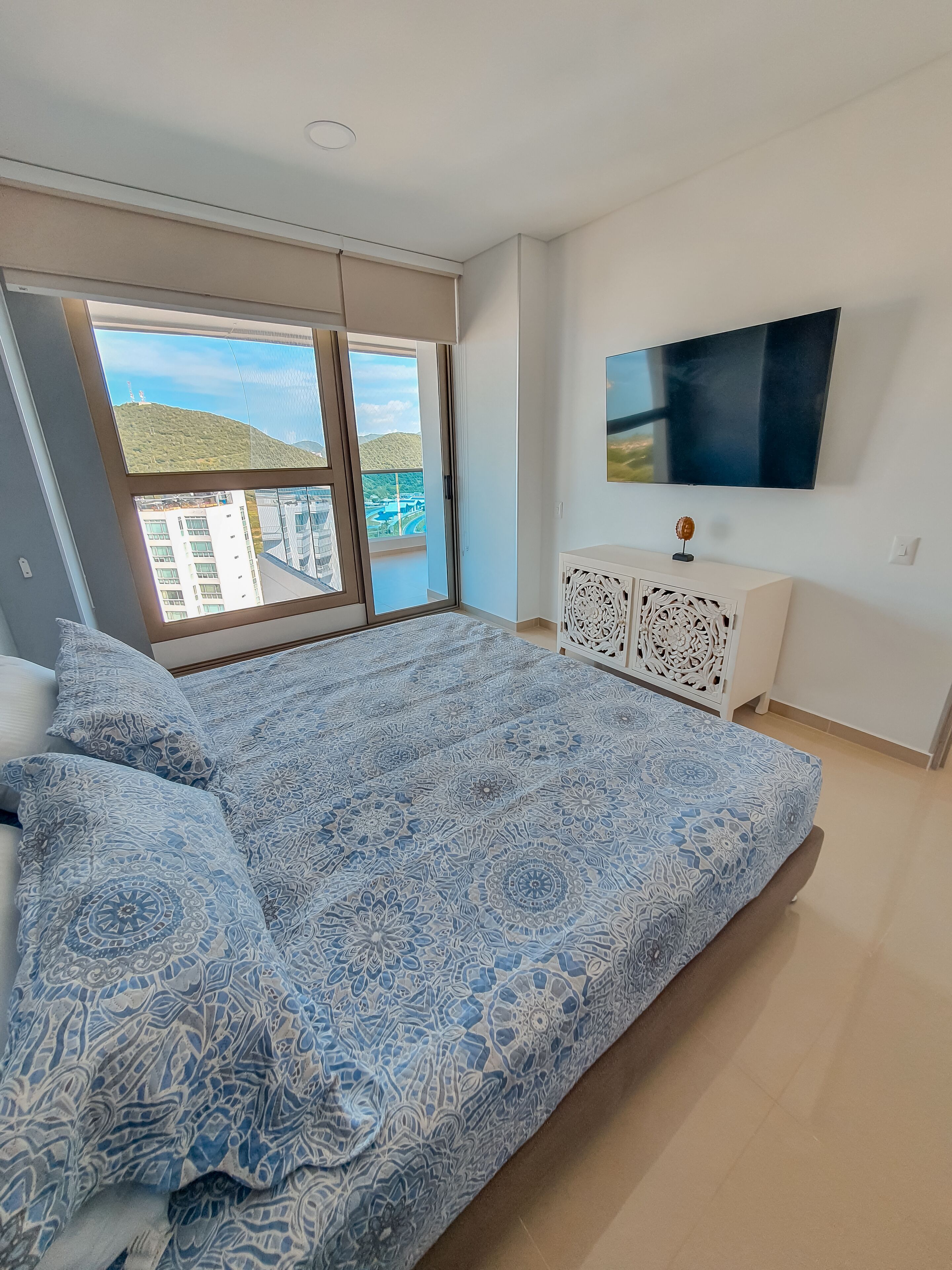A bedroom with a large bed, a TV on the wall, and a view of a cityscape through the window.