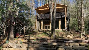 Un chalet rustique avec un escalier en pierre menant à une terrasse en bois, entouré d’arbres et d’un mur de pierres.