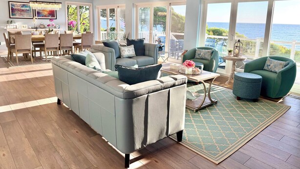 A spacious living room with a large sofa, a dining area, and a balcony with a view of the ocean.