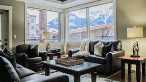 A living room with a sofa, armchairs, a coffee table, and a fireplace. Large windows offer a view of mountains and a building. - Upgraded Cozy 3 Bdrm Corner Suite, Great Mountain Views, Resort and Spa (Canmore)