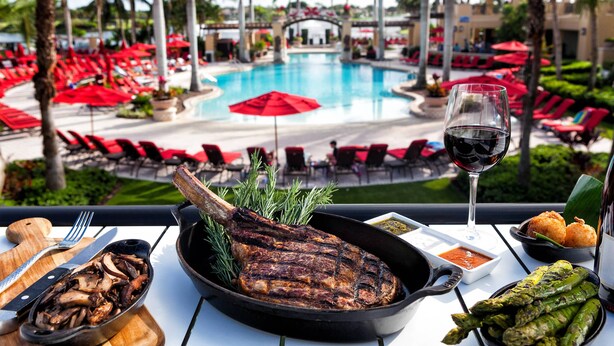 Una mesa de comedor con un filete a la parrilla, espárragos y una copa de vino tinto, con vistas a una zona de piscina con sombrillas y sillones de descanso rojos.