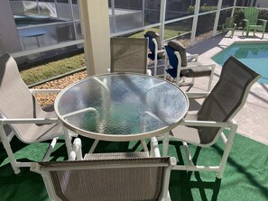 A covered outdoor seating area with a glass table and wicker chairs, overlooking a swimming pool.